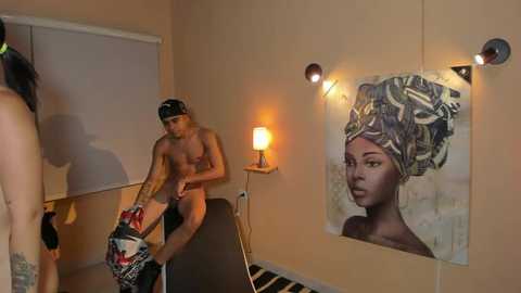 Media: Video of a nude man with a black cap, standing next to a nude woman with a headband, in a beige room with a large, colorful painting of a woman and a lamp.