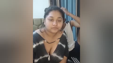 Media: Video of a young South Asian woman with medium brown skin, wearing a low-cut, black and white striped dress, sitting in a living room with a beige couch and blue curtains.