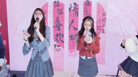 Media: Video of two Asian women, one in a striped jacket and skirt, the other in a red sweater and grey skirt, singing into microphones, against pink backdrop with traditional Chinese characters.