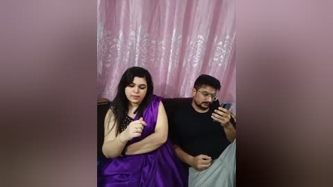 Media: Video of a woman in a purple sari and a man in a black shirt and grey pants, both looking at their phones, against a pink, floral-patterned backdrop.