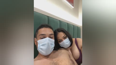 Media: A video of a shirtless man with a light complexion, wearing a face mask, and a topless woman with dark hair and large breasts, also in a face mask, standing closely in a dimly lit room with a green padded wall.