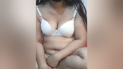 Media: Video of a woman with medium skin tone and long dark hair, wearing a white lace bra and matching panties, sitting on a red chair with her legs crossed, revealing stretch marks on her abdomen.