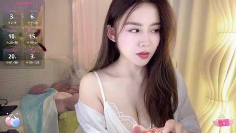 Media: Video of an East Asian woman with long brown hair, wearing a white lace bra, sitting on a bed, looking thoughtful. Background includes a calendar, yellow curtains, and a nightstand.