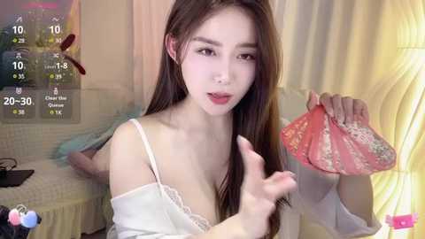 Media: Video of an East Asian woman with long brown hair, wearing a white lace camisole, holding a pink floral purse. She's indoors with a yellow curtain and a cityscape background.