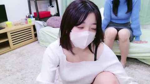 Media: Video of an Asian woman with short black hair, wearing a white face mask and sweater, kneeling on a beige carpet in a simple bedroom with a light green bed and wooden shelves.