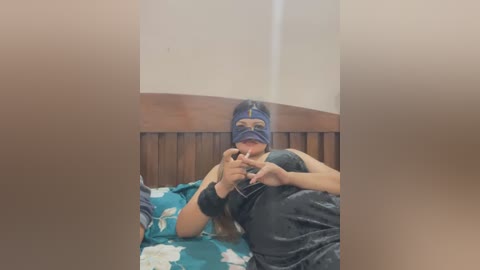 Media: Video of a woman in a blue mask, lying on a bed with a teal floral sheet, wearing a black tank top, holding a cigarette.