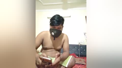 Media: Video of a shirtless man with a black mask, holding a smartphone, sitting on a bed with red sheets in a simple, dimly lit room.