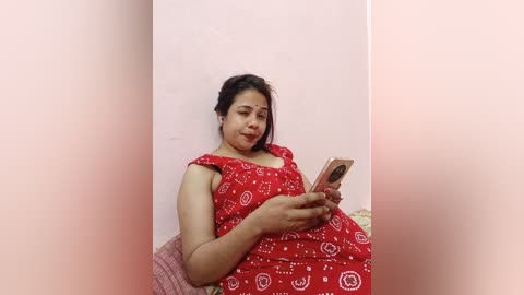 Media: Video of a South Asian woman with medium brown skin, wearing a red sari with white floral patterns, seated on a couch, holding a smartphone, against a light pink wall.