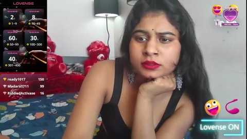 Media: Video of a South Asian woman with long black hair and fair skin, wearing a black sleeveless top, looking contemplative. Background shows a red teddy bear and a colorful bedspread.