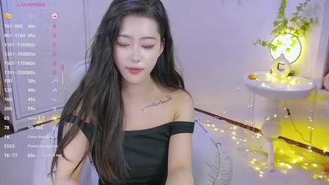 Media: Video of an Asian woman with long black hair, wearing a black off-shoulder dress, in a softly lit room with fairy lights and a round table.