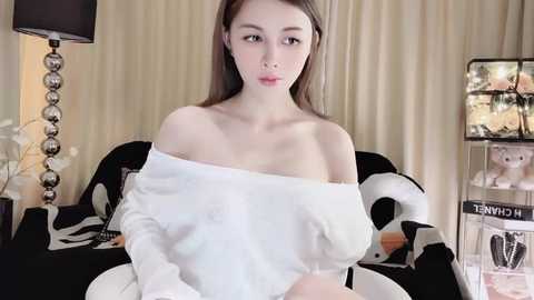 Media: Video of a young, fair-skinned woman with straight, light brown hair, wearing a white off-shoulder top, sitting on a black couch. The background features beige curtains, a lamp, and a toy display.