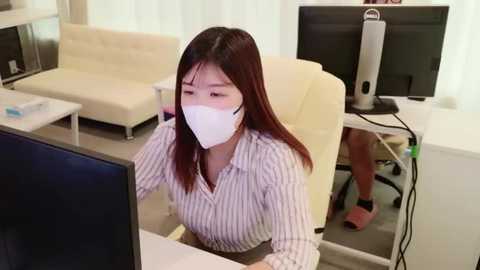 Media: Video of an East Asian woman in a white pinstriped blouse, white mask, and long brown hair, working at a desk in a modern office with white furniture, computer, and monitor.