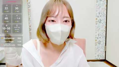 Media: A video of an Asian woman with shoulder-length blonde hair and a white mask, wearing a white top. Background shows a digital clock and a colorful floral patterned wall.