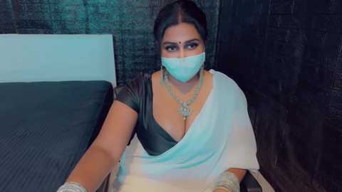 Media: Video of a South Asian woman with dark skin, wearing a white sari, black blouse, gold jewelry, and a face mask, seated on a bed against a dark brick wall.