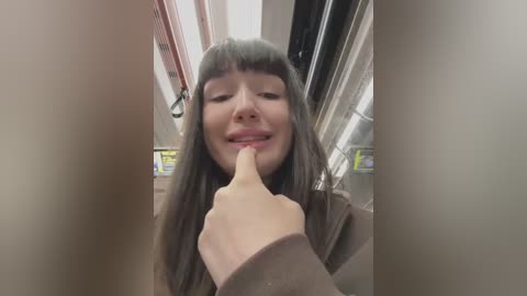 Media: Video of a smiling Asian woman with long, straight black hair, wearing a brown sweater, holding a finger to her lips, in a subway train with fluorescent lighting and advertisements.