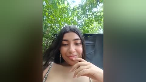 Media: Video of a young Latina woman with long dark hair, light skin, and a petite frame, wearing a leopard-print top, standing in a lush green garden. She's smiling softly, with her fingers gently touching her lips.