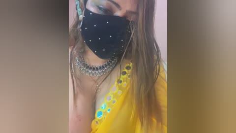 Media: Video of a woman with light skin, wearing a black face mask with silver studs, yellow blouse with blue floral embroidery, and a silver necklace, set against a blurred, neutral background.