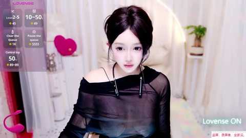 Media: Video of a young East Asian woman with pale skin, dark hair, and large hoop earrings, wearing a sheer black top, sitting in a cozy, white room.