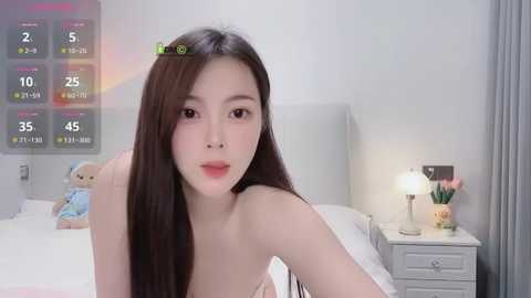 Media: A video of an East Asian woman with long, straight black hair, wearing a pink top, sitting on a bed in a modern, light-colored bedroom.