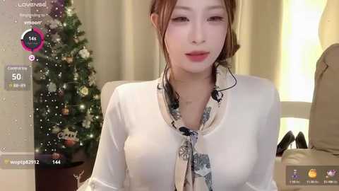 Media: Video of an Asian woman with light skin, brown hair, wearing a white blouse with a floral scarf, smiling. Background features a decorated Christmas tree and a beige curtain.