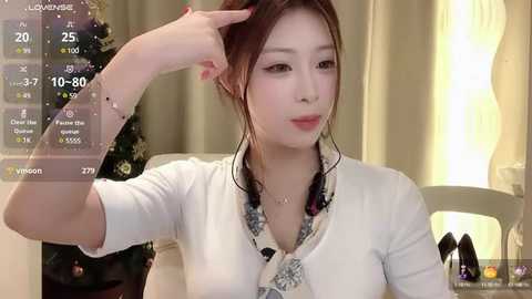 Media: A video of an East Asian woman with fair skin, brown hair, and pink lipstick, adjusting her hair. She wears a white cardigan and floral blouse, seated at a table with a Christmas tree and a glowing lamp in the background.