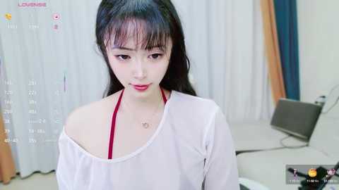 Media: Video of an East Asian woman with long black hair and fair skin, wearing a white off-shoulder top, standing in a modern, minimalist room with white curtains and a laptop.