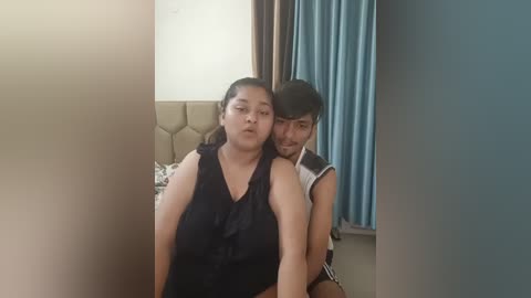 Media: A video of an overweight woman with dark hair, wearing a black top, and a slender man with short black hair, wearing a white and black sleeveless shirt, embracing each other in a cozy bedroom with beige headboard and blue curtains.