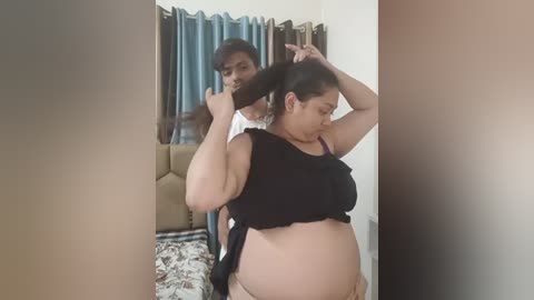 Media: Video of a pregnant woman in a black crop top, tying her hair back while standing in a dimly-lit living room with beige curtains and a patterned blanket.