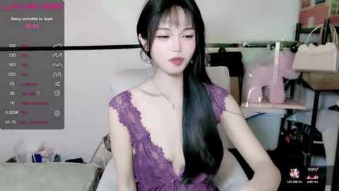 Media: Video of an East Asian woman with long black hair, fair skin, and full lips, wearing a purple lace dress, sitting in a cluttered room with a shelf of clothes in the background.