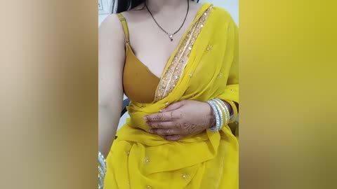 Media: Video of a woman in a mustard yellow sari with intricate gold embroidery, holding her stomach, wearing a gold necklace, and multiple bangles on her wrist.