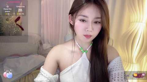 Media: A video of an East Asian woman with long, straight brown hair, wearing a white off-shoulder top and a green beaded necklace. She's in a dimly lit room with soft, pastel-colored curtains and a bed.