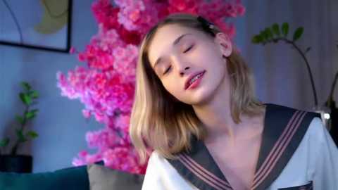 Media: Video of a young Caucasian woman with blonde hair, wearing a Japanese schoolgirl sailor uniform, smiling softly. Background features a pink floral arrangement and a potted plant.