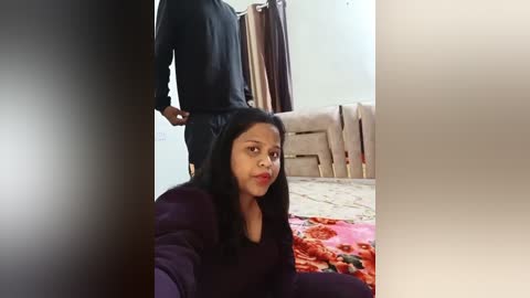 Media: Video of a South Asian woman with long black hair, wearing a purple sweater, sitting on a red blanket in a beige room with a bed, brown curtains, and a man standing behind her.