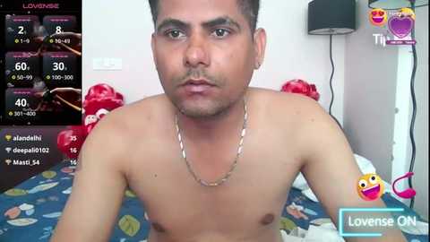 Media: Video of a shirtless man with medium skin tone, short dark hair, and a necklace, sitting on a bed with colorful bedding. Background includes a phone displaying social media statistics and a heart emoji.