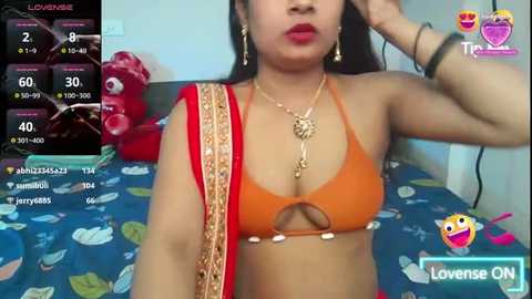 Media: A video of an Indian woman with medium skin tone, wearing a bright orange bikini top with a cutout design and a red sari draped over her shoulder, posing in a bedroom with a blue bedspread.