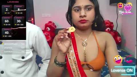 Media: Video of a South Asian woman in an orange bikini, holding a red sari, in a bedroom with teddy bears, wearing gold jewelry.