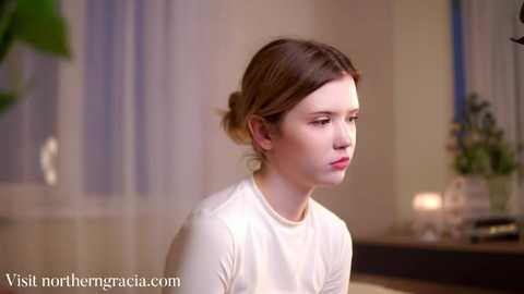 Media: Video of a young Caucasian girl with fair skin and brown hair in a simple white top, seated indoors with a blurred background featuring a potted plant and a window.