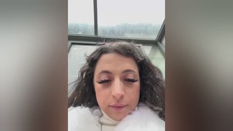 Media: Video of a woman with curly brown hair and closed eyes, wearing a white fur coat, standing in front of a large window with a blurred forest view outside.