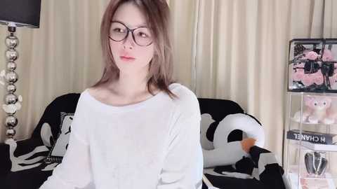 Media: A video of a fair-skinned Asian woman with straight, shoulder-length brown hair, wearing black-rimmed glasses, a white sweater, and sitting on a black couch adorned with plush toys, including a white whale.