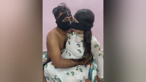 Media: Video of a shirtless Black man with a black mask and a South Asian woman with long black hair, both wearing white floral-patterned dresses, embracing closely.