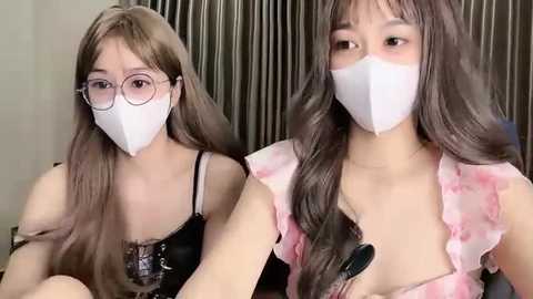 Media: Video of two young Asian women with long, wavy brown hair, wearing white face masks, glasses, and revealing black lace lingerie. They are indoors with a gray curtain background.