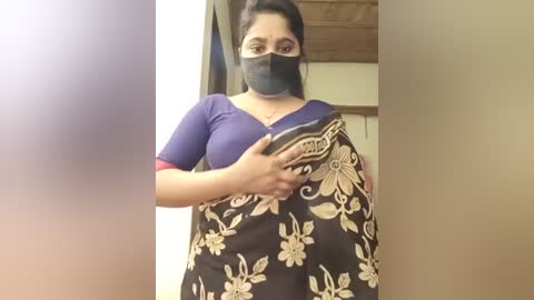 Media: Video of a young woman with medium skin tone, wearing a black face mask, blue blouse, and floral-patterned sari, holding a smartphone, in a room with wooden ceiling and beige walls.