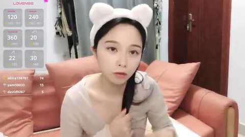 Media: Video of an Asian woman with pale skin, wearing a white bear headband and a light grey sweater, seated on a pink couch in a modern living room.