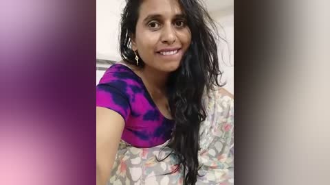 Media: A video of a smiling South Asian woman with long black hair and medium skin tone, wearing a vibrant purple and black tie-dye top and a floral saree, set against a blurred background.