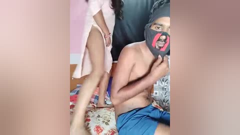 Media: Video of a woman in pink lingerie and a man in a black mask and blue shorts, standing and sitting on a patterned bed in a bedroom.