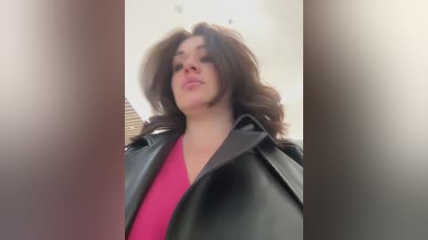 Media: Video of a woman with voluminous, wavy brown hair, wearing a black leather jacket over a pink shirt, standing in a modern, bright room with beige walls and a large window with horizontal blinds.