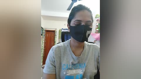 Media: Video of a young woman with dark hair in a ponytail, wearing a gray t-shirt with \"California\" text, black face mask, and standing indoors. Background includes a wooden door, patterned curtains, and a ceiling fan.