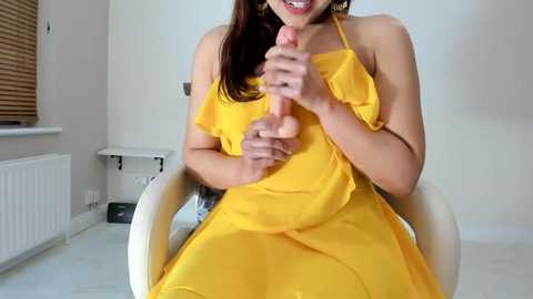 Media: Video of a smiling, light-skinned woman with long brown hair, wearing a yellow dress, holding a large, erect dildo in her hands, sitting in a white chair against a plain white wall and window with blinds.