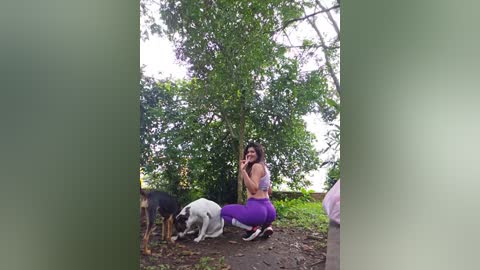 Media: Video of a young woman with long dark hair, wearing a purple sports bra and leggings, kneeling on a dirt path in a lush, green park, petting a white and brown dog.