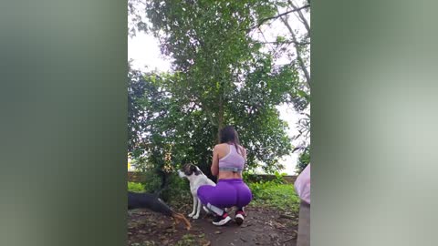 Media: Video of a woman with dark hair, wearing a white sports bra and purple leggings, crouched in a forest, petting a black and white dog.